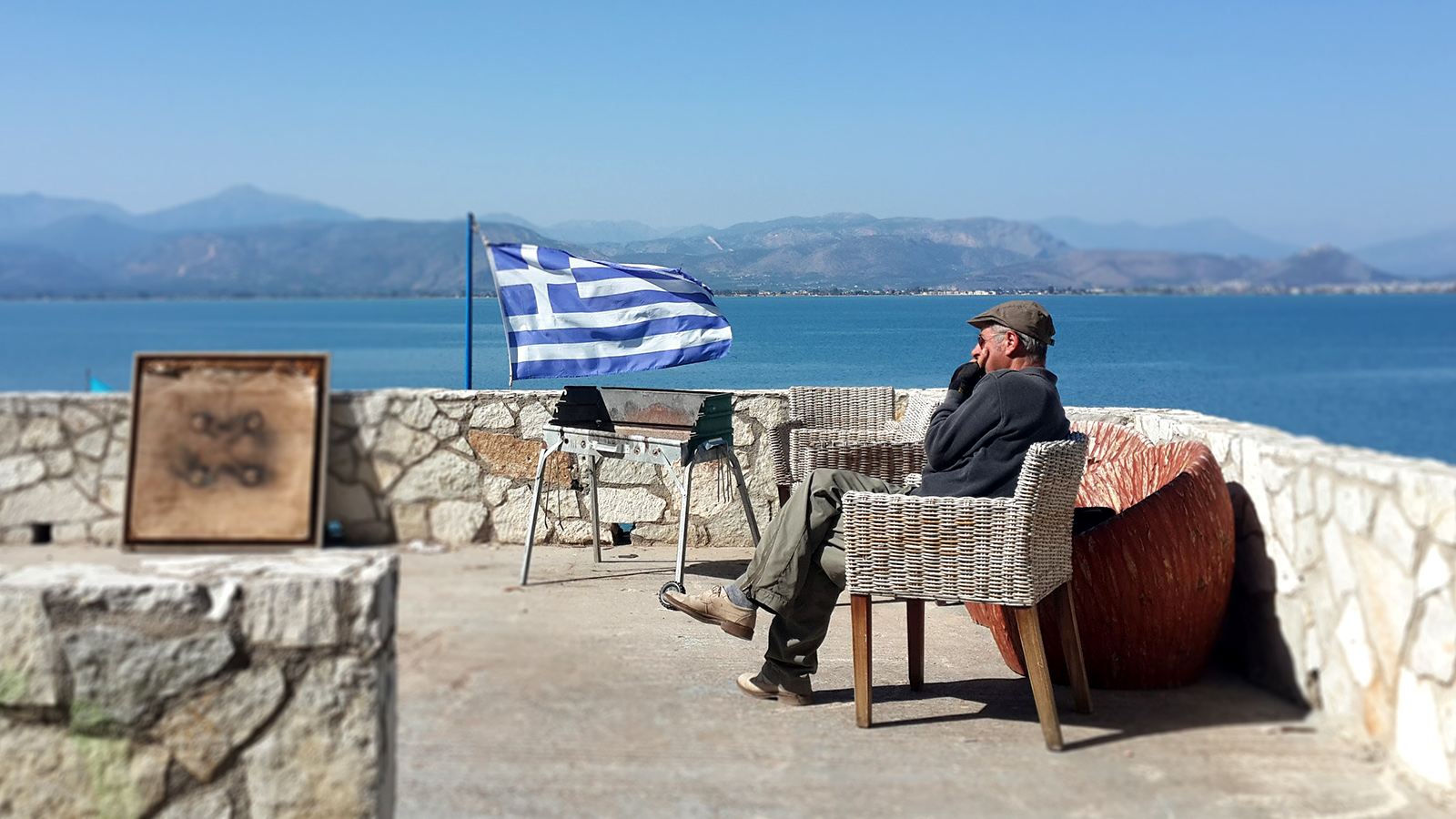 יווני זקן מביט לנוף כשברקע דגל יוון. (צילום: רז רותם).