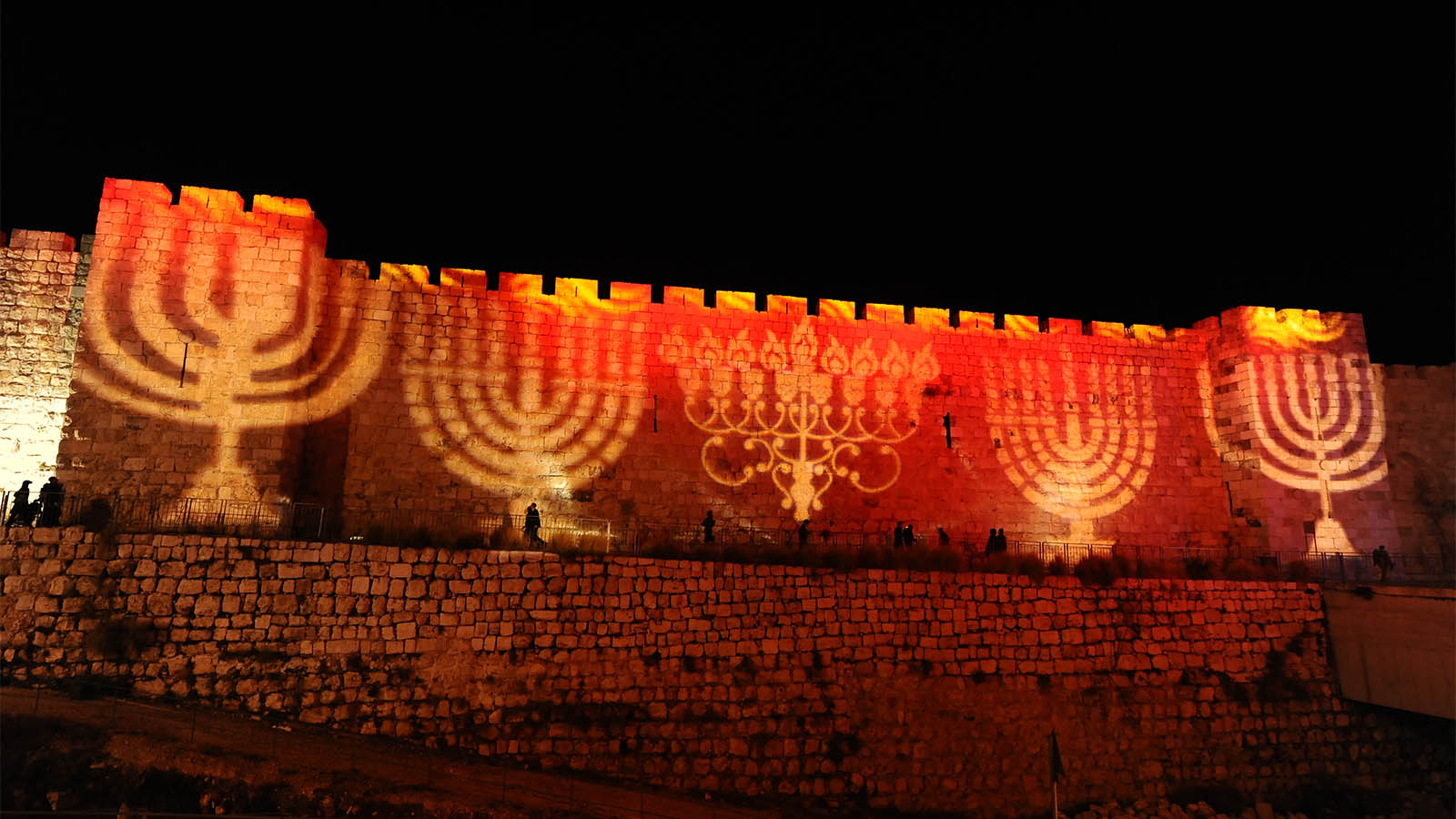 Pepole walk past the walls of Jerusalem's Old City illuminated with projection showing Menorah and Dreidels during the Jewish holiday of Hanukkah. December 24, 2016 (Photograph by Mendy Hechtman/Flash90)