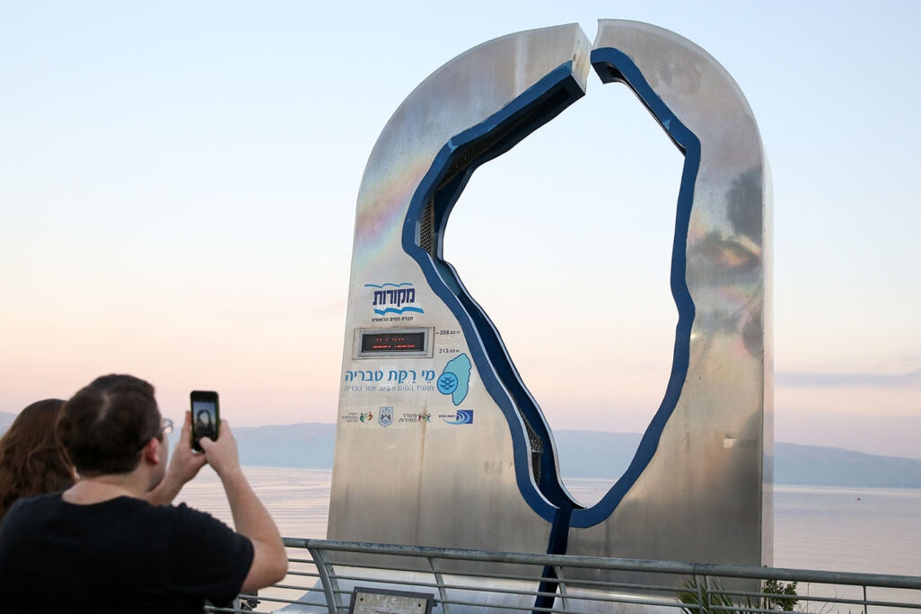 Visitors standing near a sign displaying the Kinneret water level on the beach promenade of the Northern Israeli city of Tiberias, right by the Kinneret lake on March 18, 2019. (Photograph: David Cohen/Flash90)