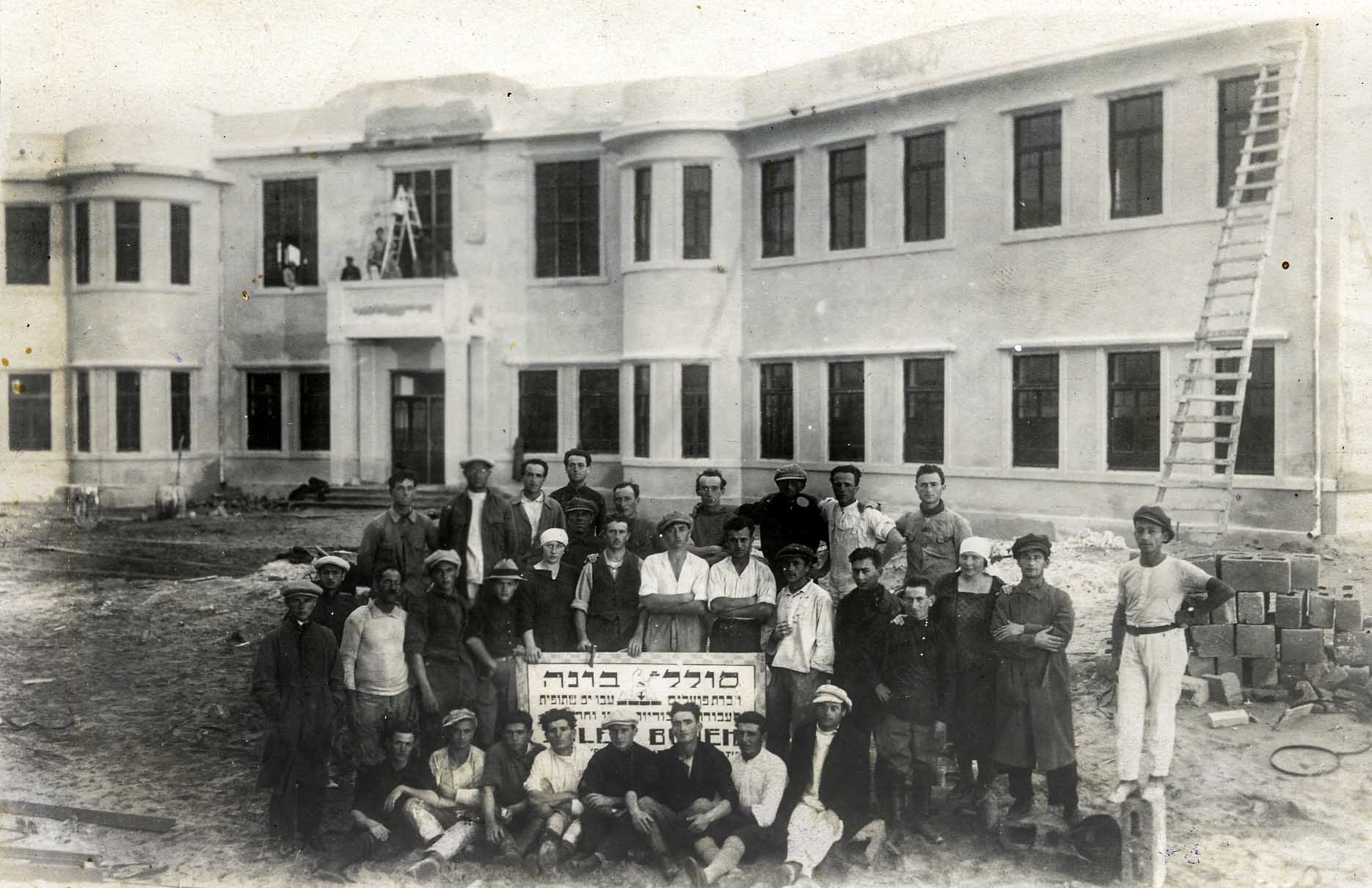 קבוצת פועלי הבניין 'יסוד' בתל אביב, על רקע בית הספר תל נורדוי בגמר בנייתו. בית הספר נבנה על ידי סולל בונה (מתוך אוסף מכון לבון)