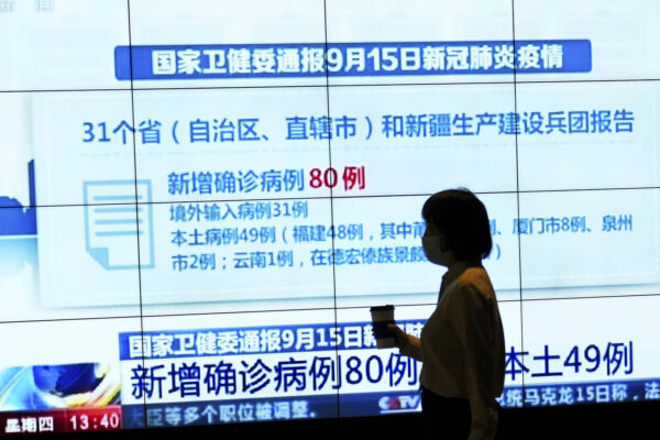 אישה עם מסכה במרכז קניות בבייג'ינג עוברת ליד מסך טלוויזיה עליו מוקרנים נתוני תחלואת הקורונה בסין. (צילום: AP Photo/Andy Wong)