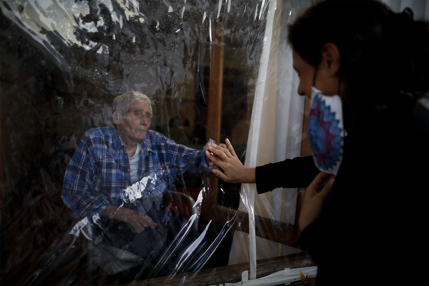 מעון לקשישים בארגנטינה. ויקטור טריפיאנה בן ה-86 מושיט יד לגעת בידי כלתו, סילביה פרננדס סוטו, כשהוא מבודד על ידי יריעת פלסטיק עקב הקורונה. 4 באפריל 2021 (צילום: AP Photo/Natacha Pisarenko)