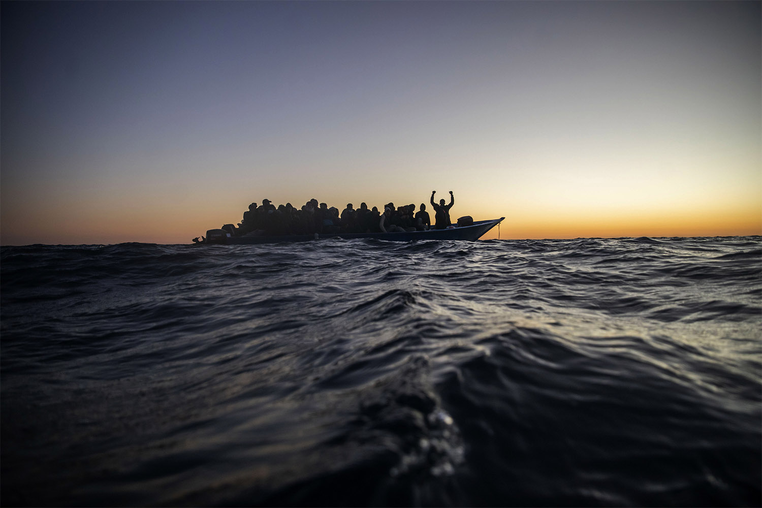 מהגרים ופליטים בני לאומים אפריקאיים שונים מחכים לסיוע על סירת עץ צפופה. עובדי סיוע של העמותה הספרדית ׳זרועות פתוחות׳ מתקרבים אליהם בים התיכון, במים הבינלאומיים, במרחק של 196 ק״מ מהחוף הלובי. 12 בפברואר 2021 (צילום: AP Photo/Bruno Thevenin)