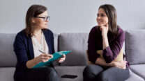 Social worker, psychologist talking to young woman in office. Women sitting on couch, counselor making notes, listening to patient. Psychology, mental health, social difficulties of young people