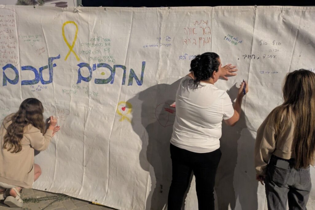 מבוגרים וילדים כותבים על בד גדול מסרים לחטופים לקראת שובם הצפוי, עצרת קהילת ניר עוז בכרמי גת, 11 באוקטובר 2025 (צילום: קרן שמש)