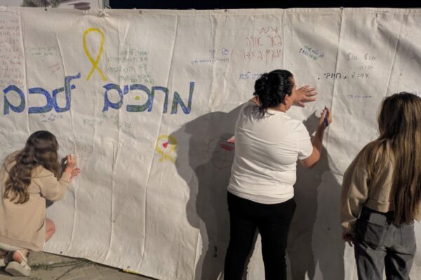 מבוגרים וילדים כותבים על בד גדול מסרים לחטופים לקראת שובם הצפוי, עצרת קהילת ניר עוז בכרמי גת, 11 באוקטובר 2025 (צילום: קרן שמש)