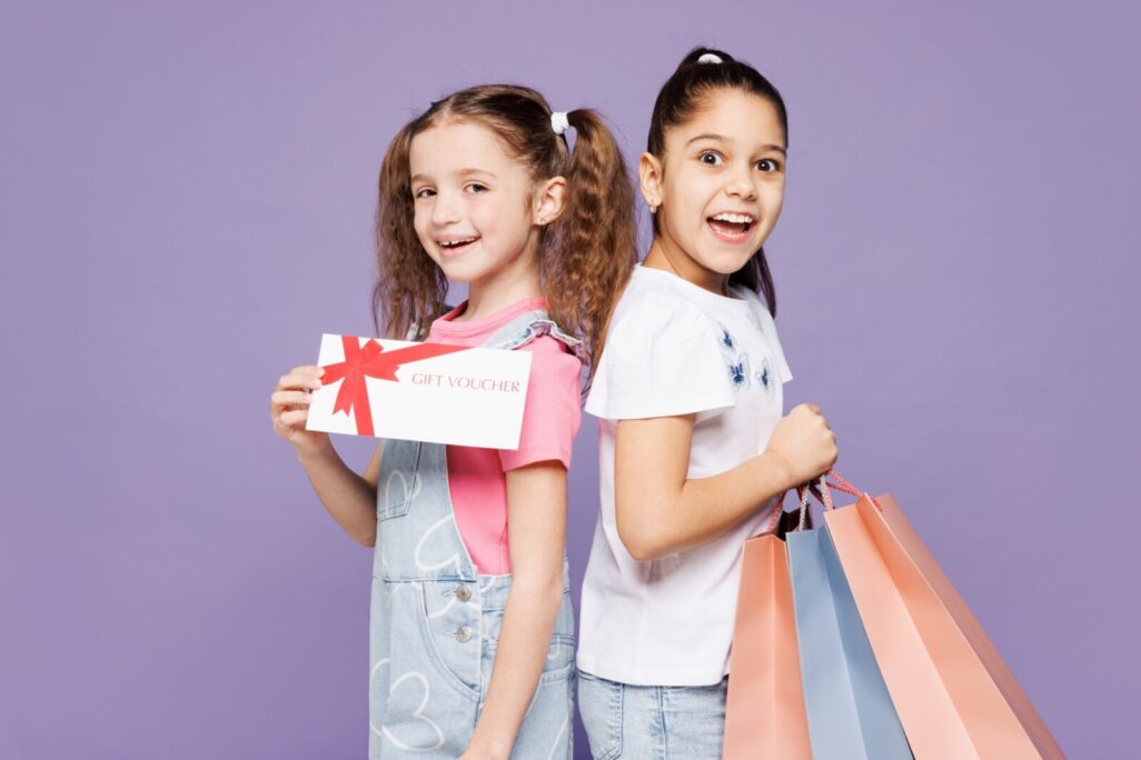 שתי חברות בנות 9-10, לובשות בגדים יומיומיים יחד, אוחזות בקניות, שקיות נייר, כרטיס שובר, מבודד על רקע סגול פשוט. מבצעי יום שישי השחור, קונספט של יום קנייה (צילום: Shutterstock)